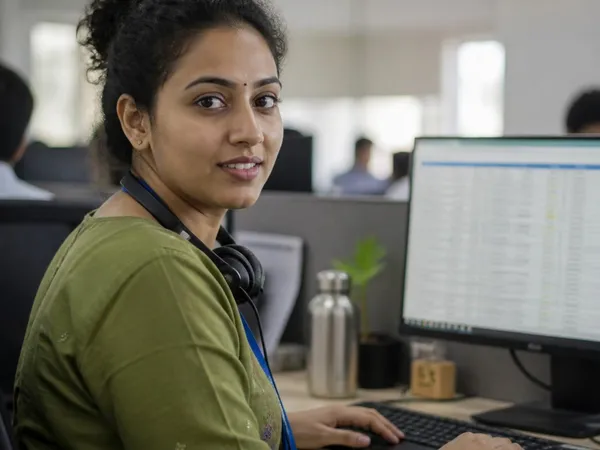 A woman in an open office with a headset around her neck, looking at the camera, a monitor with a records table beside her.
